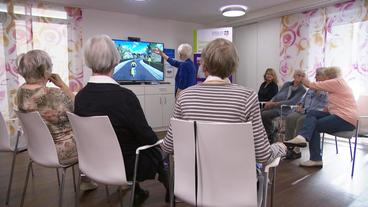Eine ältere Frau steht vor einem Bildschirm und spielt ein Computerspiel. Eine Gruppe Seniorinnen sitzt auf Stühlen im Halbkreis um sie herum.