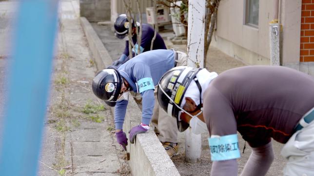 Drei Personen mit Helm reinigen gebückt eine niedrige Betonmauer