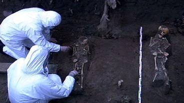 Zwei Wissenschaftler in weißem Schutzanzug vermessen ein geöffnetes Grab mit Skelett.