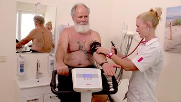 Ein Mann sitz auf einem Ergometer, eine Arzthelferin misst den Blutdruck.