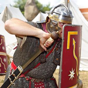 Ein Soldat im Kettenhemd steht in Angriffsstellung sein Schwert ziehend vor weissen Zelten eines Roemerlagers