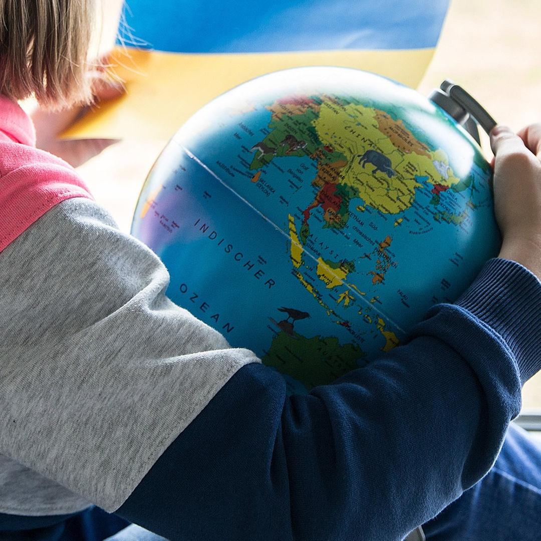 Ein Kind sitz vor dem Fenster mit einer Weltkugel Globus und hält die Flagge der Ukraine in der Hand.