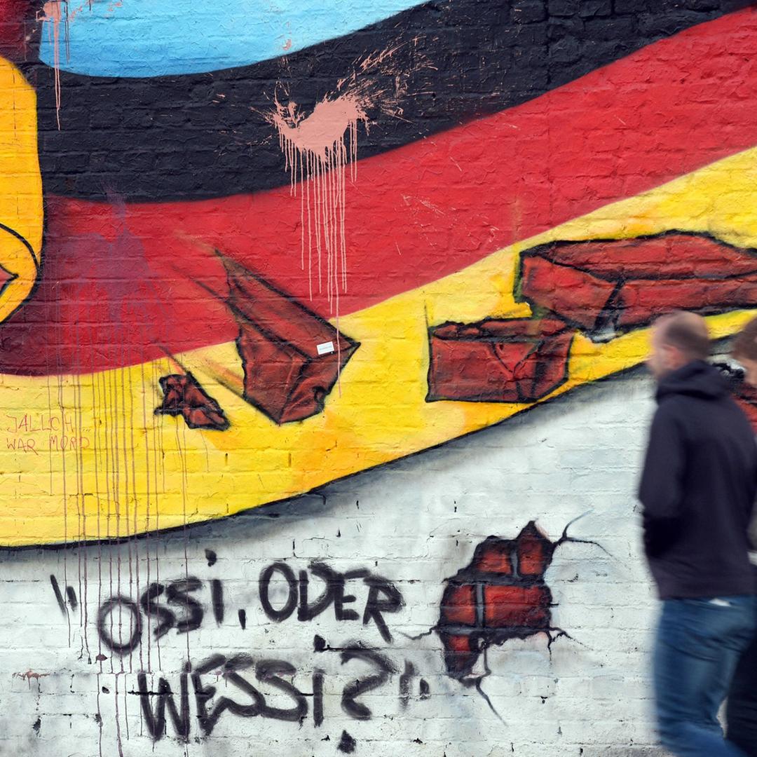 Passanten gehen am 02.10.2015 in Berlin an an einem Wandbild von Caspar Kirchner mit der deutschen Nationalflagge und dem Schriftzug "Ossi oder Wessi? vorbei.