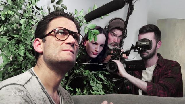 Christian Ehring mit Brille, daneben eine Zimmerpflanze, hinter der sich ein Kamerateam versteckt.