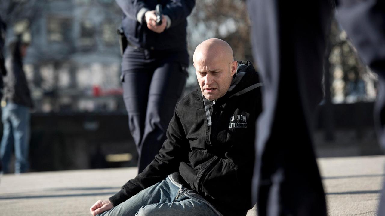 Frank liegt am Boden, Polizisten laufen auf ihn zu.