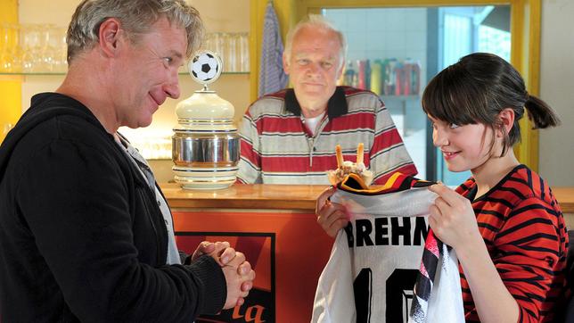 Die fußballbegeisterte Charlie (Annika und Francesca Bolkart) strahlt über ein Geschenk ihres Papas Alex (Jörg Schüttauf); auch Opa Willy (Peter Franke) freuts…