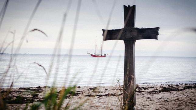 Ein Kreuz am Strand. Im Hintergrund ist das Schiff zu sehen.