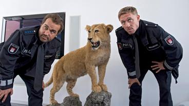 Bild: ARD/Thorsten Jander Falk Lohmanns ganzer Stolz – ein von ihm selbst geschossener Löwe. Paul Dänning (Jens Münchow) und Piet Wellbrook (Peter Fieseler) sehen sich in ihren Vorurteilen gegen den Finanzexperten voll bestätigt.