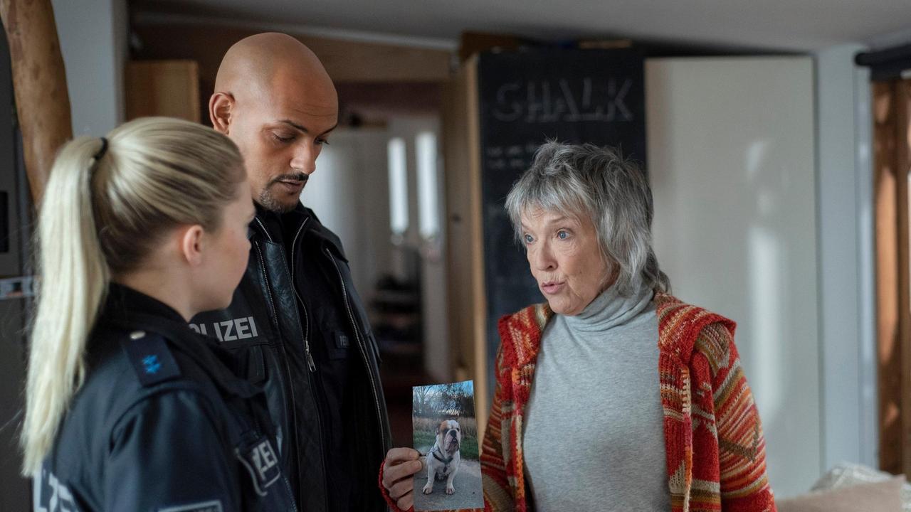 Monika Böckhold (Eva Kryll) zeigt Lukas Petersen (Patrick Abozen) und Jessy Jahnke (Farina Flebbe) ein Foto ihrer verschwundenen englischen Bulldogge Morpheus. Die beiden Polizisten sollen sie suchen.