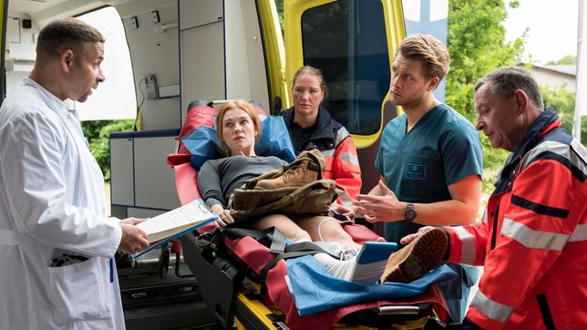 Bild: ARD/Jens-Ulrich Koch Ariane Bischof (Franziska Brandmeier, 2.v.l. mit Komparsin, h.) wird direkt vom Flughafen in das Johannes-Thal-Klinikum eingeliefert. Dr. Moreau (Mike Adler) und Mikko Rantala (Luan Gummich, 2.v.r. mit Komparse) nehmen sie in der Notaufnahme auf.
