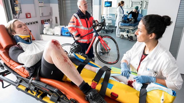 Bild: ARD/Jens Ulrich Koch Der Rennrad-Sportler Andreas Lott (Felix Kruttke, l. mit Komparse, h.) wird mit einem offenen Bruch in die Notaufnahme eingeliefert. Dr. Leyla Sherbaz (Sanam Afrashteh) beginnt mit der Erstuntersuchung.