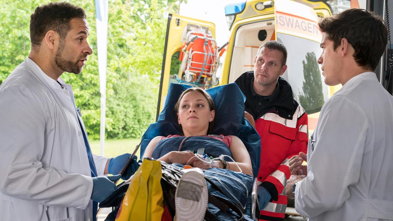 Die Buchverkäuferin Sarah Hahn (Mathilde Bundschuh, M. mit Komparse, h.) ist gestürzt und kommt mit einer Knöchelverletzung ins JTK. Dr. Matteo Moreau (Mike Adler) und Ben Ahlbeck (Philipp Danne) nehmen sie auf.