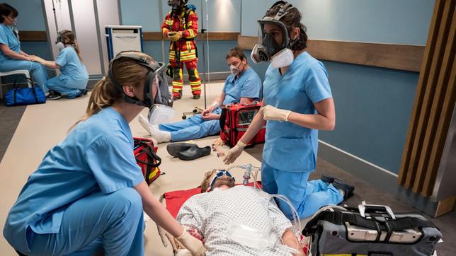 Bild: ARD/Jens-Ulrich Koch Feuerwehreinsatz im Johannes-Thal-Klinikum! Unter Einsatz von Atemmasken wird Linus Haas (Nikolay Sidorenko) nach einem Herzstillstand, von Rebecca Krieger (Milena Straube) und Julia Berger (Mirka Pigulla) versorgt (mit Kompars:innen und (Christian Beermann, h.).