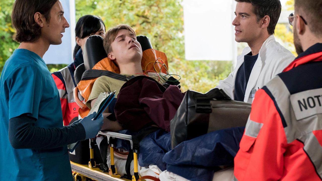 Ivo (Jakob D'Aprile) und Dr. Ben Ahlbeck (Philipp Danne, r. mit Komparse) nehmen gemeinsam den stark alkoholisierten Teenager Georg Freytag (Oliver Szerkus, M. mit Komparsin, h.) im JTK auf.