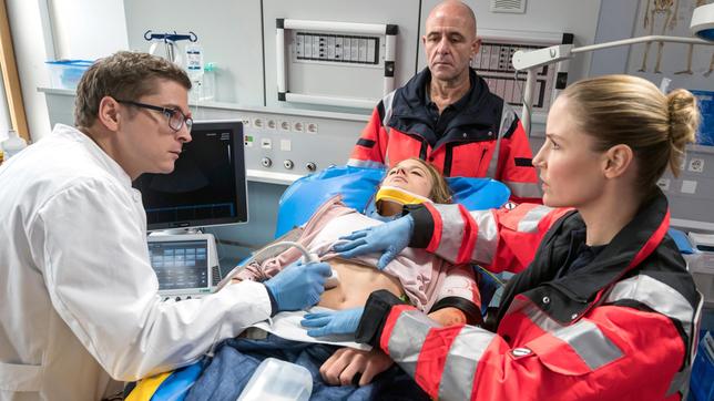 Bild: ARD/Jens Ulrich Koch Julia Berger (Mirka Pigulla) kennt die jungen Patientin Vanessa Nimmrich (Sinha Marie Gierke, M. mit Komparse, h.) aus dem Kinderwunschzentrum. Elias Bähr (Stefan Ruppe) erfährt von Julia, dass Vanessa bereits neun gescheiterte Versuche, schwanger zu werden, hinter sich hat.