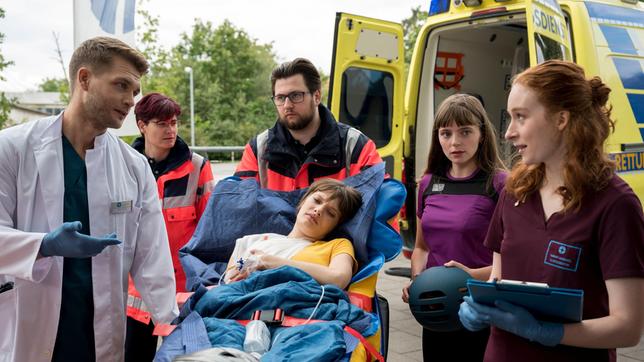 Bild: ARD/Jens-Ulrich Koch Nele Bader (Sarah Schmidt) wird in Begleitung ihrer Freundin Bianka Schneider (Annemarie Hörold) in das Johannes-Thal-Klinikum eingeliefert. Mikko Rantala (Luan Gummich) und Tamar Hummel (Linda Kummer) nehmen die Patientin vom Rettungsdienst (Komparsen, h.) entgegen.