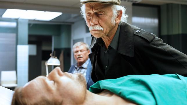 Dr. Jamm (Michael Kors) ist ermordet worden. Edwin Bremer (Tilo Prückner) mustert die Leiche, Kollege Günter Hoffmann (Wolfgang Winkler, im HG) hält sich im Hintergrund.