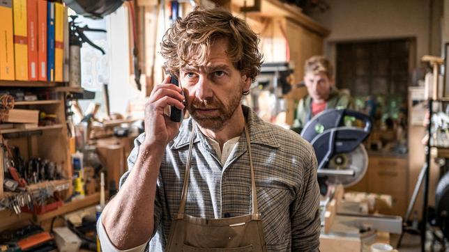 Schreinermeister Sebald Wimmer (Andreas Nickl) telefoniert im Beisein seines Sohnes Moritz (Lorenzo Nedis Walcher) mit seiner der Bank.