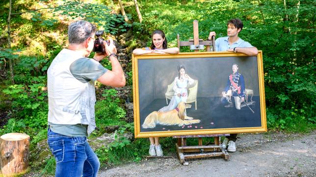 Joshua (Julian Schneider) und Denise (Helen Barke) posieren mit dem Bild von Dora und George vor dem Fotograf (Komparse).