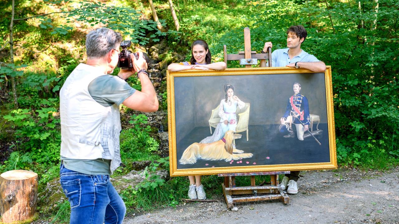 Joshua (Julian Schneider) und Denise (Helen Barke) posieren mit dem Bild von Dora und George vor dem Fotograf (Komparse).