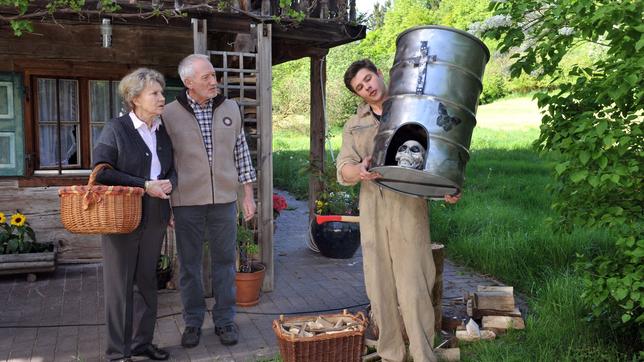 Alfons (Sepp Schauer, M.) und Hildegard (Antje Hagen, l.) sind fassungslos über die mit Totenköpfen bemalte Regentonne, die ihnen Jonas (Sebastian Fritz, r.) als sein Kunstwerk "Memento Mori" präsentiert